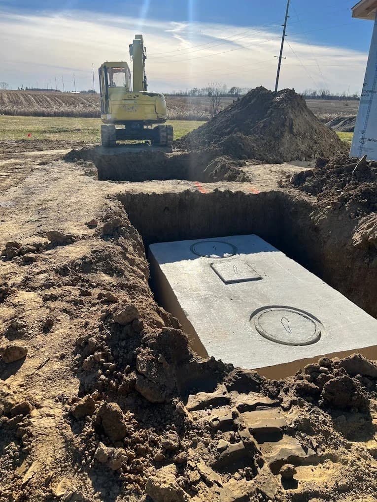 Concrete septic tank set level in an open excavation with a mini excavator and spoil pile behind it on a Champaign County property