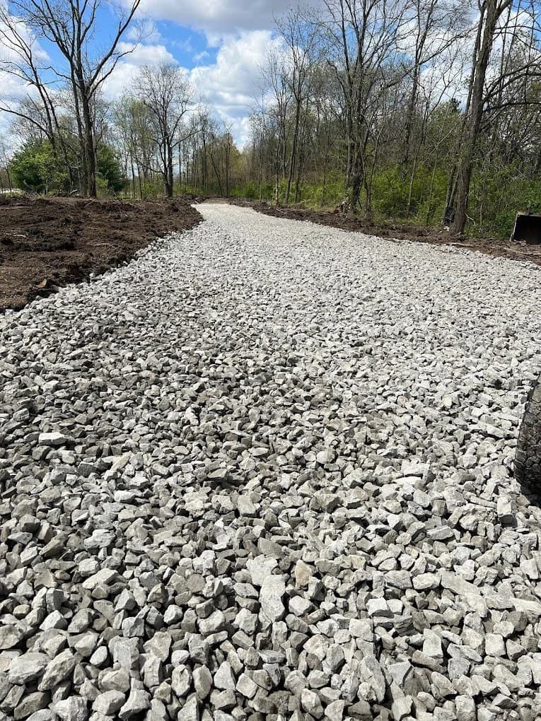 Freshly placed white limestone gravel lane cut through a wooded lot with graded dirt shoulders on both sides